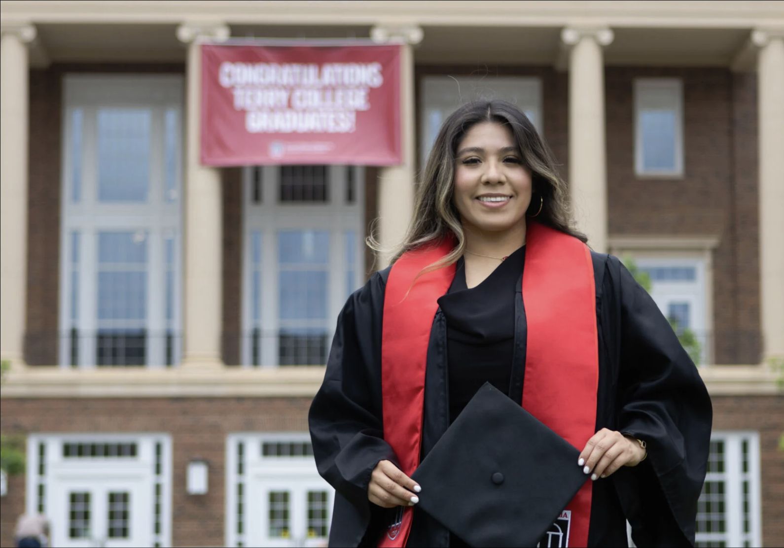 A young woman wears her graduation robe and stands proudly in front of a University building